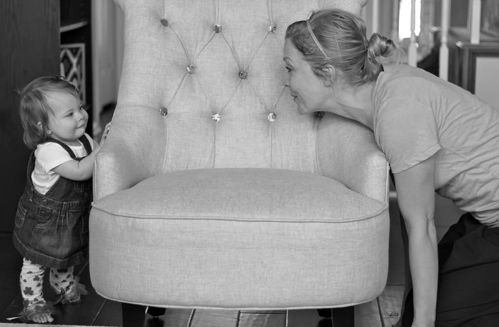 Nine month old infant playing peek a boo with mother behind chair